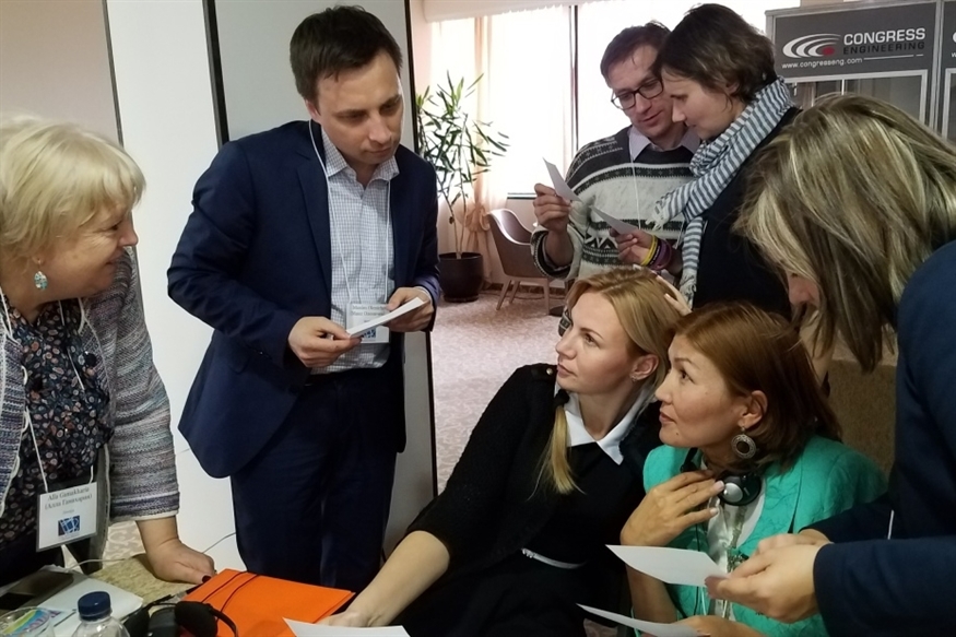 [ai] A group of six people engaged in a discussion around a table, with two women seated and four individuals standing, holding papers. The setting appears to be a business or conference environment.